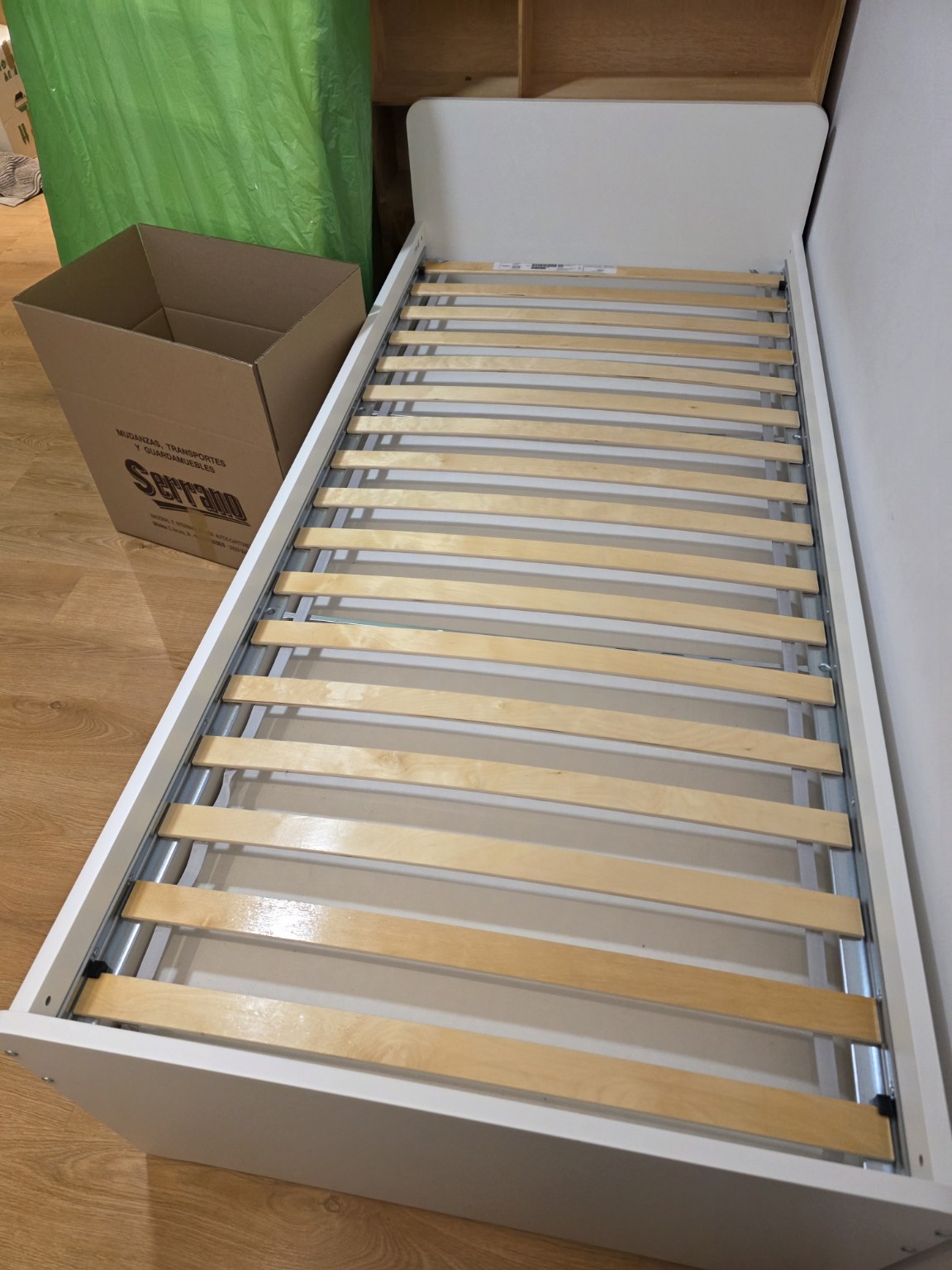 Somier con lamas de madera junto a una caja de Mudanzas Serrano y piezas de una cama